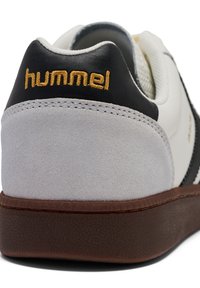 Sneaker bianco e grigio con accenti neri. Presenta un tallone in camoscio, suola marrone texturizzata e logo "hummel" dorato sul retro.