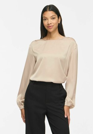 Femme aux longs cheveux foncés portant un chemisier beige à manches longues et un pantalon noir, debout devant un fond clair uni.