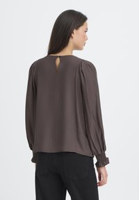 Brun langærmet bluse med gathered ærmer, nøglehul-lukning i ryggen og struktureret stof. Båret med sorte bukser.
