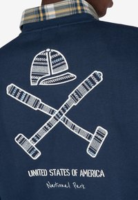 Camisa navy por detrás con palos de croquet cruzados y parche de casco encima del texto "Estados Unidos de América Parque Nacional"; persona con cuello a cuadros visible.