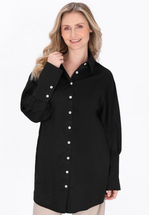 Femme souriante aux cheveux blonds ondulés portant une chemise noire à manches longues avec des boutons blancs, debout devant un fond blanc.