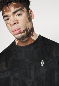 Černé sportovní tričko s texturovaným, vzorovaným designem. Má kulatý výstřih a malé bílé logo na hrudi. Hladká tkanina.