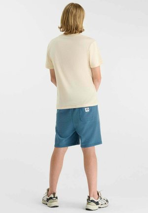 Jeune personne aux cheveux blonds vue de dos, portant un t-shirt beige, un short bleu et des chaussures de sport blanches sur un fond uni.