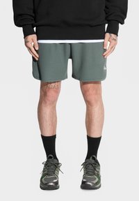 Zielone szorty o fakturze waffle z kontrastującym wykończeniem, w połączeniu z czarnymi skarpetami sięgającymi do kostek i butami sportowymi w kamuflażowym wzorze. Na górze czarna bluzka.