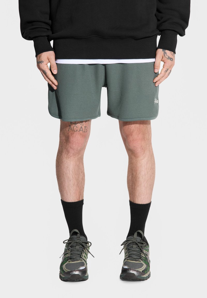 Zielone szorty o fakturze waffle z kontrastującym wykończeniem, w połączeniu z czarnymi skarpetami sięgającymi do kostek i butami sportowymi w kamuflażowym wzorze. Na górze czarna bluzka.