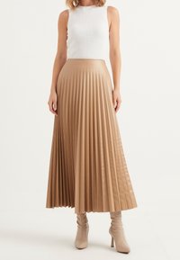 Fuktad midikjol i ljusbrun konstläder, kombinerad med en vit ribbad ärmlös topp. Kjolen har vertikala veck och en svävande design.