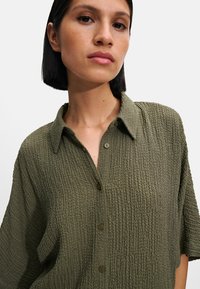 Oliwkowa, teksturowana koszula z kołnierzykiem rozłożonym, z przodu zapinana na guziki, z krótkim rękawem. Tkanina ma pomarszczony wygląd.