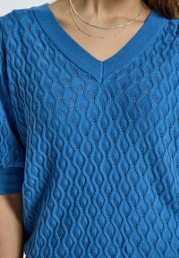 Modrý pletený svetr s výstřihem do V, texturovaným vlnitým vzorem a krátkými rukávy. Materiál vypadá lehký a prodyšný.