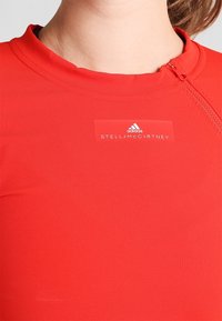 Červený sportovní top s vysokým límcem, se zipovým uzávěrem. Branding zahrnuje bílé štítky "STELLA McCARTNEY" a "adidas". Hladká textura.
