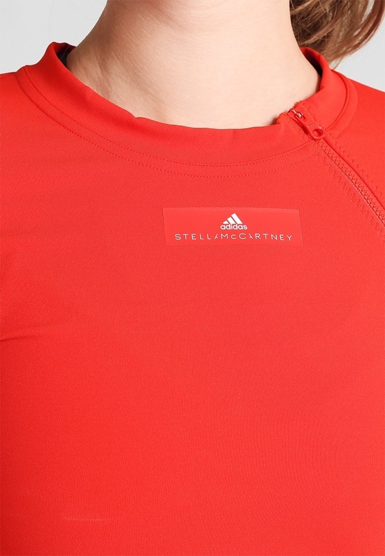 Červený sportovní top s vysokým límcem, se zipovým uzávěrem. Branding zahrnuje bílé štítky "STELLA McCARTNEY" a "adidas". Hladká textura.
