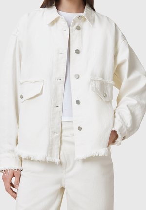 Kvinde iført en cremefarvet denimjakke med frynsekant, metaltryknapper og to frontlommer med klap, stylet med matchende cremefarvede bukser.