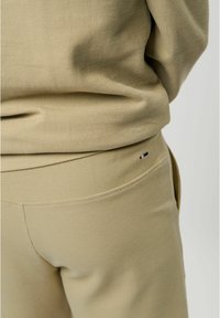 Gros plan d'une personne portant un pantalon beige texturé et un haut assorti, montrant le détail du tissu et un petit logo brodé près de la poche.