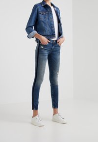Kvinna stående med händerna i fickorna, iklädd en blå denimjacka, slitna skinny jeans med svarta detaljer, vita sneakers och vit topp.
