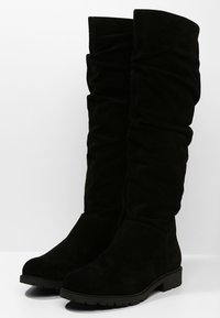 Musta mokkanahkainen polvipituinen saapas, joka on löysästi muotoiltu, litteällä pohjalla ja vähäisellä varustuksella. Pehmeä rakenne, jossa on sileä viimeistely.