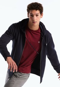 Haut à zip bleu marine avec cordons de serrage et poches avant, superposé sur un T-shirt bordeaux. Un pantalon gris complète l'ensemble. Texture lisse.