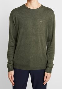 Mies, joka pukeutuu oliivinvihreään pitkähihaiseen pyöreäkaulaiseen neuleeseen, jossa on pieni brodeerattu logo rinnassa, yhdistettynä tummiin housuihin.