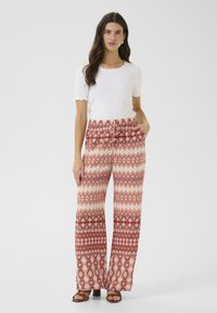 Pantalons larges à motifs en crème et rouge avec des designs complexes ; associés à un haut blanc à manches courtes en côte et des sandales marron.