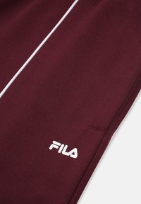 Tessuto tecnico bordeaux con piping bianco. Logo Fila in bianco sul lato sinistro in basso. Texture liscia, design affinato, dettagli di cucitura puliti.