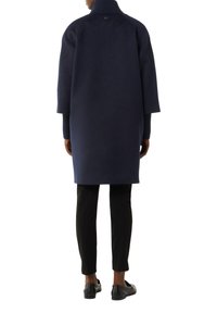 Marineblauer Wollmantel mit hohem Kragen, überschnittenen Schultern und verdecktem Knopfdetails auf der Rückseite. Kombiniert mit schlanken schwarzen Hosen und schwarzen Slippern.