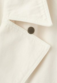 Witte denimkleding in close-up met een spits kraagje en een metalen knoop met de opdruk "Established 1984 King Denim".