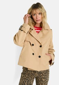 Femme blonde portant un trench-coat beige à double boutonnage, une chemise à rayures rouges et blanches, et un pantalon en imprimé léopard, posant avec la main près du visage.