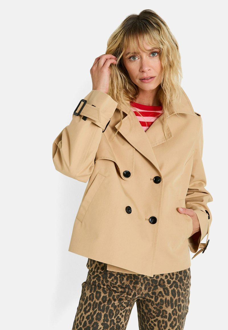 Femme blonde portant un trench-coat beige à double boutonnage, une chemise à rayures rouges et blanches, et un pantalon en imprimé léopard, posant avec la main près du visage.