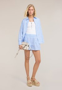 Joven mujer con camisa de rayas azul claro y pantalones cortos con pequeños detalles florales, camiseta blanca sin mangas, sandalias de plataforma y llevando un bolso blanco cruzado.