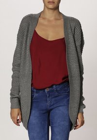 Woman wearing a loose gray knitted cardigan over a red sleeveless top and blue jeans, standing with arms relaxed by her sides.