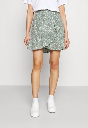 Grüner Wickelrock mit Punkten und einem gerafften Saum, mit seitlicher Drapierung und strukturiertem Stoff. Kombiniert mit weißen Turnschuhen und Socken.