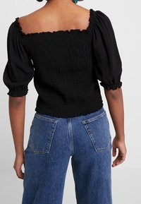 Černý nabíraný top s balónovými rukávy a čtvercovým výstřihem, kombinovaný s modrými džíny s vysokým pasem a velkými zadními kapsami.