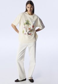 Crème katoenen t-shirt met kleurrijke tropische graphic, gecombineerd met off-white wijde broek en zwarte platte schoenen.