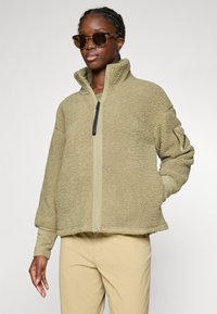 Donna che indossa una giacca in sherpa di colore sabbia con zip frontale, maniche lunghe e occhiali da sole, in piedi contro uno sfondo chiaro e uniforme.