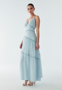 Světle modré maxi šaty s vrstveným designem, nastavitelnými tenkými ramínky a jemně texturovanou látkou s ozdobným prošíváním.
