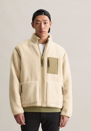 Young man wearing a beige fleece jacket with green zipper pockets and cuffs, paired with a black backward cap and dark pants.