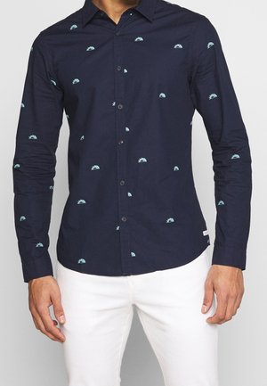 Mann trägt ein marineblaues Hemd mit hellblauen Halbkreismustern und weiße Hose, steht mit entspannten Händen.