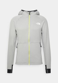 Veste à capuche gris clair avec fermeture éclair jaune sur le devant, poches latérales zippées, poignets noirs et logo blanc North Face sur la poitrine.
