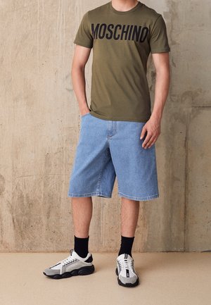 Homme portant un t-shirt Moschino vert olive, un short en jean bleu clair, des chaussettes noires et des baskets noir et blanc devant un mur beige.