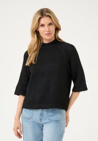 Schwarzer Strickpullover mit hohem Kragen, kurzen Ärmeln und lockerer Passform. Weiche Textur und leicht flauschige Oberfläche; passt gut zu hellblauen Jeans.