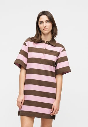 Γυναίκα που φοράει καφέ και ροζ κοντομάνικο φόρεμα πόλο με φαρδιές ρίγες, γιακά και κουμπότρυπες, στέκεται μπροστά σε μια μονόχρωμη επιφάνεια.