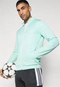 Mintgrüne Sportjacke mit vollem Reißverschluss und Stehkragen. Verfügt über zwei Taschen und einen schlanken, elastischen Stoff. Getragen mit grauen Hosen.