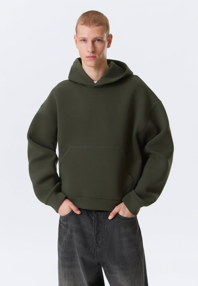 Jeune homme portant un sweat à capuche surdimensionné vert olive et un jean gris foncé, debout les mains dans les poches devant un fond uni.