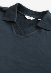 Tumed meresinine polo särk, millel on V-kaeluse disain, pehme puuvillane materjal ja kraega kaelus. Sildil on kirjas "NEXT." Sile tekstuur, minimaalne detail.