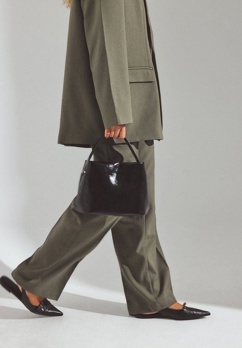 Oseba v olivno zeleni obleki hodi, drži sijočo črno torbico in nosi črne čevlje z ostrimi konicami brez zavezovanja.