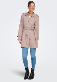 Trench-coat rose clair avec une taille ceinturée, des boutons sur le devant et des épaulettes, associé à un jean skinny bleu et des bottines noires.