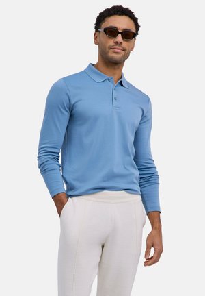 Uomo che indossa occhiali da sole scuri, polo a maniche lunghe azzurro chiaro e pantaloni chiari, in piedi con una mano in tasca.