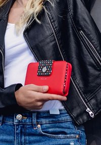 Red wallet made of leather, featuring a patterned flap with multicolored dots and a silver button, held against a black leather jacket.