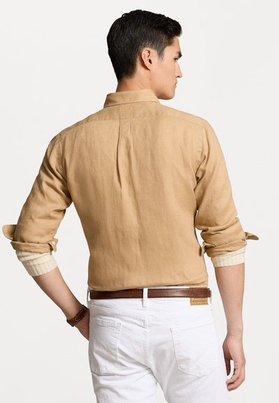 Chemise en lin beige avec des manches retroussées et des pattes à boutons, dotée d'un col classique et d'un pli au dos, assortie à un pantalon blanc et une ceinture marron.