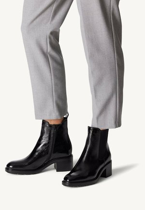 Bottines en cuir noir avec une finition brillante, dotées d'un talon carré, d'une fermeture éclair sur le côté et de panneaux élastiques sur les côtés, associées à un pantalon gris ajusté.