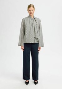 Kvinde står iført en bluse med sort og hvid skråstribede mønster, med en knyttet hals, brede sorte bukser og sorte spidse sko.