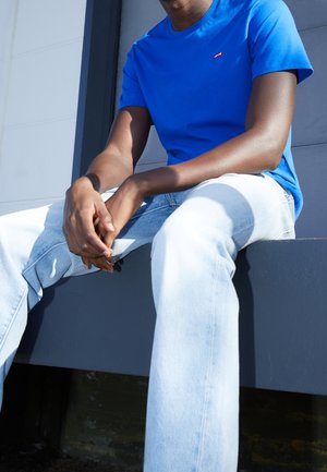 Blaues Baumwoll-T-Shirt mit kurzen Ärmeln und kleinem Logo, kombiniert mit hellblauen Denim-Jeans. Modell sitzt vor einem grauen Hintergrund.